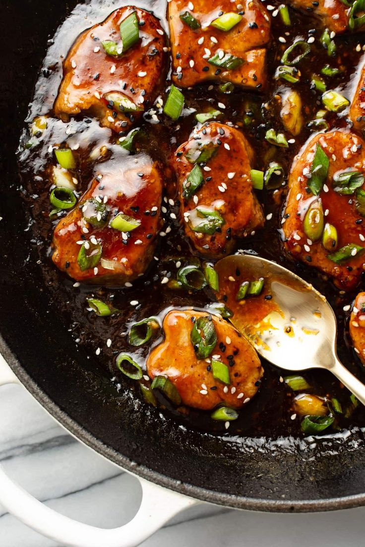 a skillet filled with chicken covered in sesame seeds and green onions next to a wooden spoon