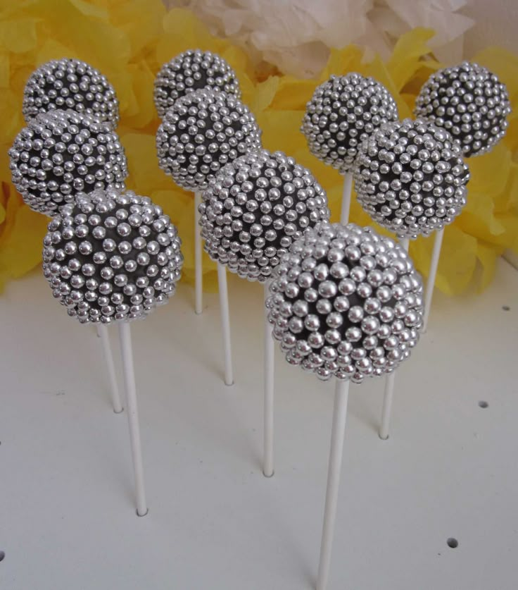 six silver cake pops sitting on top of a white table next to yellow flower petals