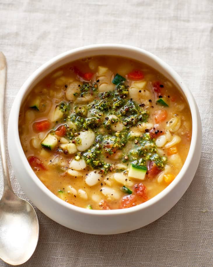 a white bowl filled with pasta and vegetables next to a spoon on top of a table