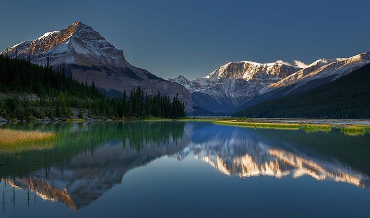 a mountain range is reflected in the still water of a lake at twilight with trees and grass around it