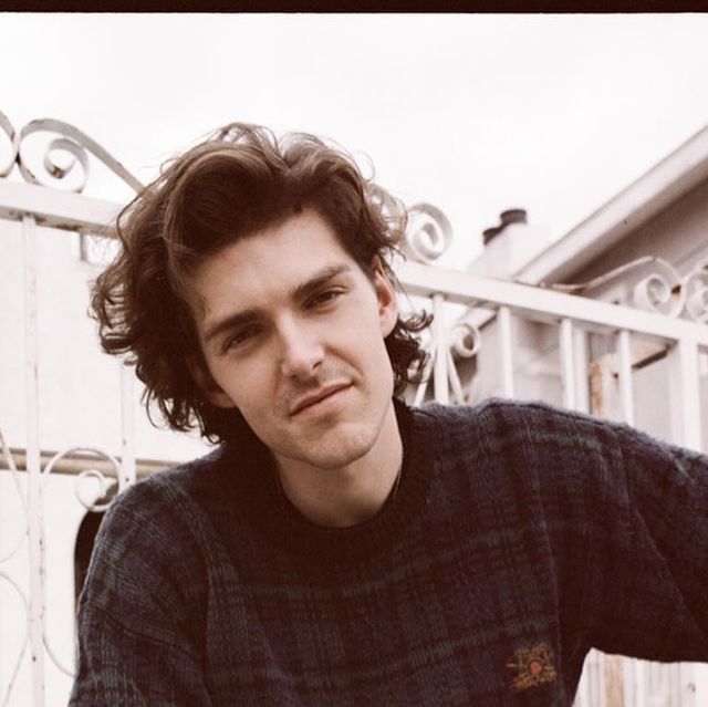 a young man with long curly hair wearing a sweater and looking at the camera while standing in front of a white fence