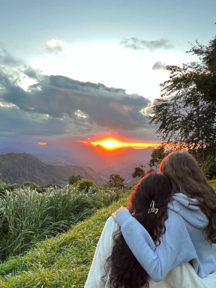 two people sitting on top of a lush green hillside under a cloudy sky at sunset