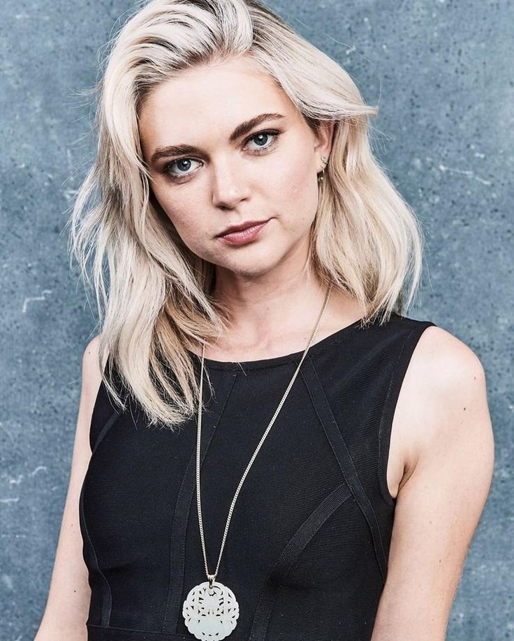 a woman with blonde hair wearing a black top and silver necklace, standing in front of a gray wall