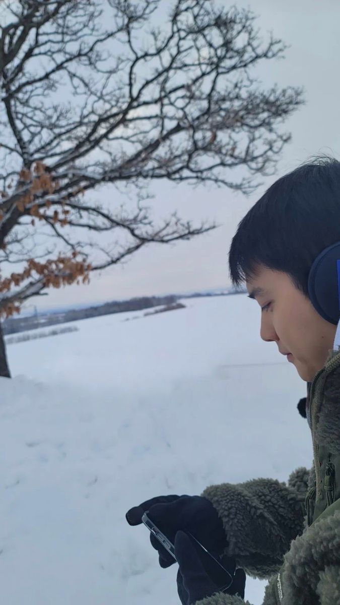 a young boy wearing ear muffs looking at his cell phone in the snow with a tree behind him