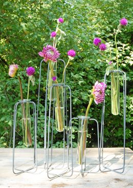 four metal vases with flowers in them sitting on a wooden table near some trees