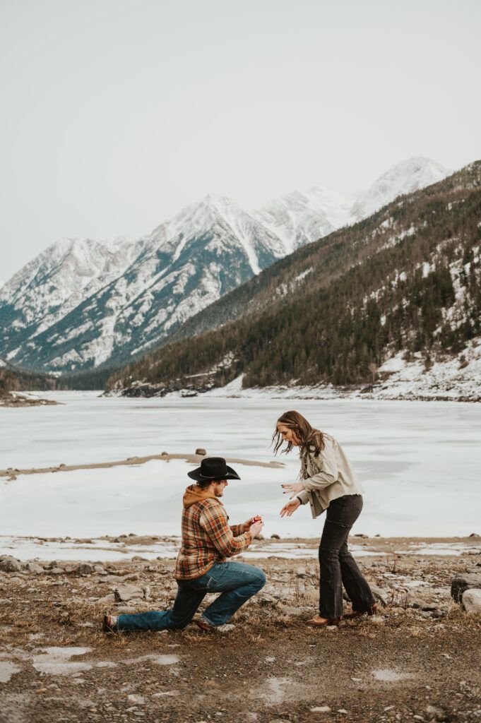 two people sitting on the ground in front of snow covered mountains, one is talking to the other