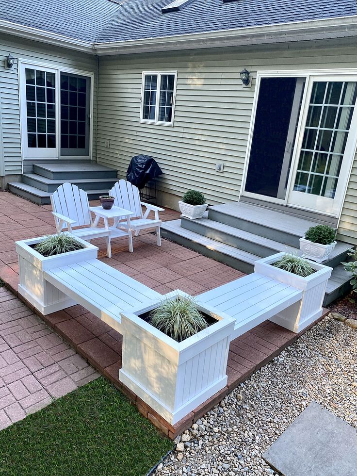 a patio with white chairs and plants on the ground next to a brick walkway in front of a house