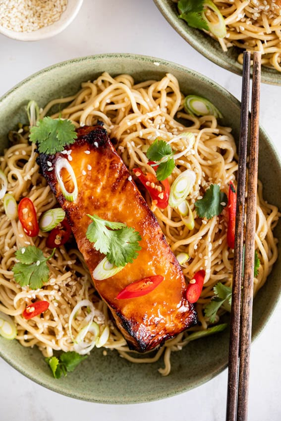 two green bowls filled with noodles and salmon on top of a white tablecloth next to chopsticks