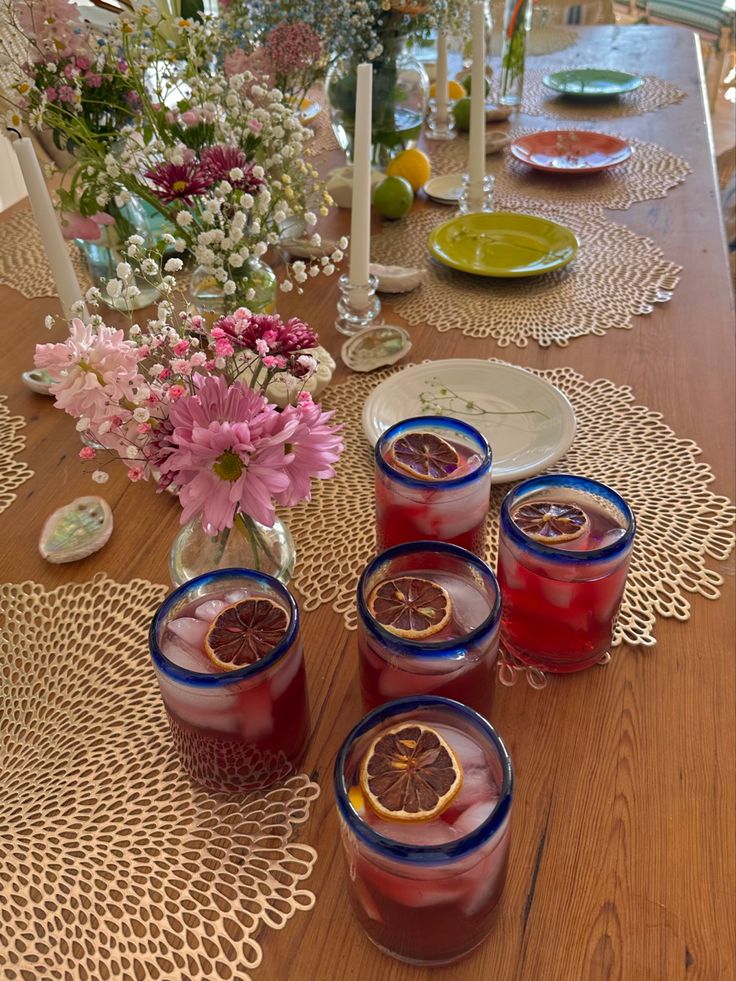 a wooden table topped with glasses filled with liquid next to plates and flowers on top of it