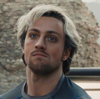 a man with white hair wearing a blue shirt and looking off into the distance while standing in front of a stone wall