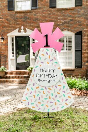 a birthday cake shaped yard staker in front of a brick house with the number one on it