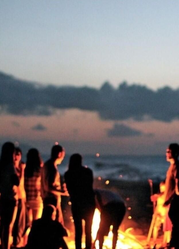 a group of people standing around a fire on the beach at night with bright lights