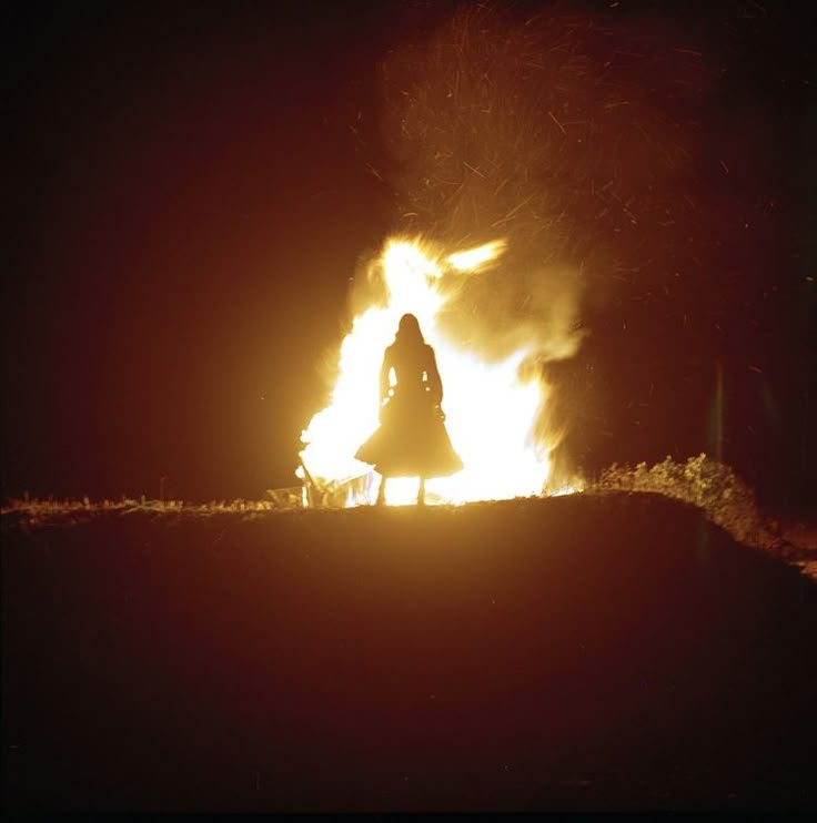 a woman standing in front of a fire with bright orange flames behind her on a dark night