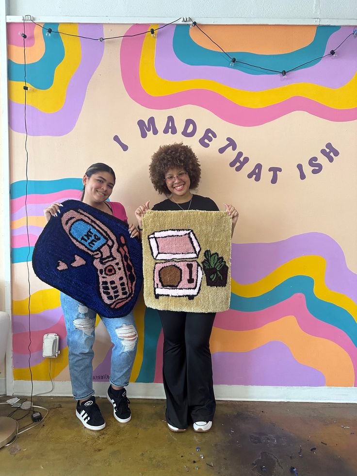 two people standing in front of a colorful wall holding up an art piece that says i madehatish