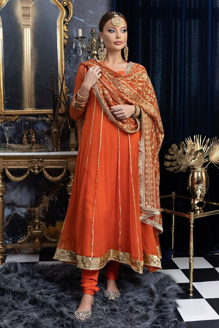 Apricot Anarkali is the dress of queens and princes. It's regal and takes craftsmanship and effort to achieve an exquisite brew of tangerine and gold. Presenting 'Apricot' the exclusive Maria Nasir design that can be your next wedding outfit for the season. The beautiful gota and tilla work, hand-cut sequins, silk threadwork in gold fits beautifully in auburn chiffon silhouette. The round neck with deep back and gorgeous tassels make this ensemble extremely gorgeous. Gold bordered panels and gold foil gota lace in the bottom brings out the style exquisitely. Paired with similar hued churidaars and heavily embroidered dupatta in chiffon fabric is all you need to turn all the heads toward you! Top: Color: Orange Fabric: Chiffon Anarkali dress with gota work Round neck 3/4 sheer sleeves with Chiffon Anarkali Dress, Orange Dress Outfits, Chiffon Anarkali, Anarkali Designs, Silk Anarkali Suits, Bridal Suits, Orange Suit, Latest Bridal Dresses, Designer Anarkali