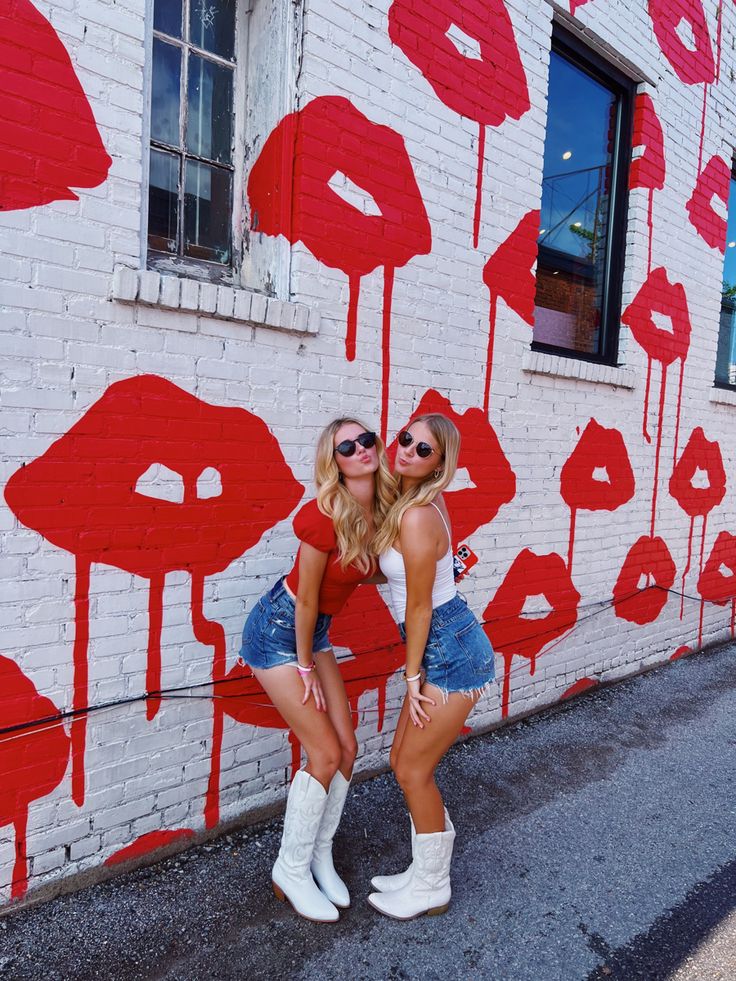 two beautiful young women standing next to each other in front of a wall with red lipstick painted on it