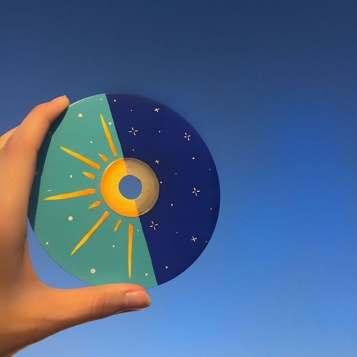 a hand holding a small disc with the sun and moon painted on it in front of a blue sky