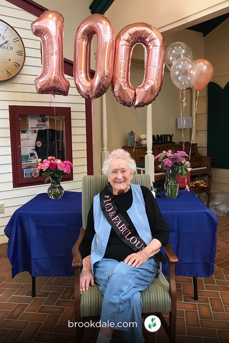 an older woman sitting in a chair with balloons and flowers on the table behind her