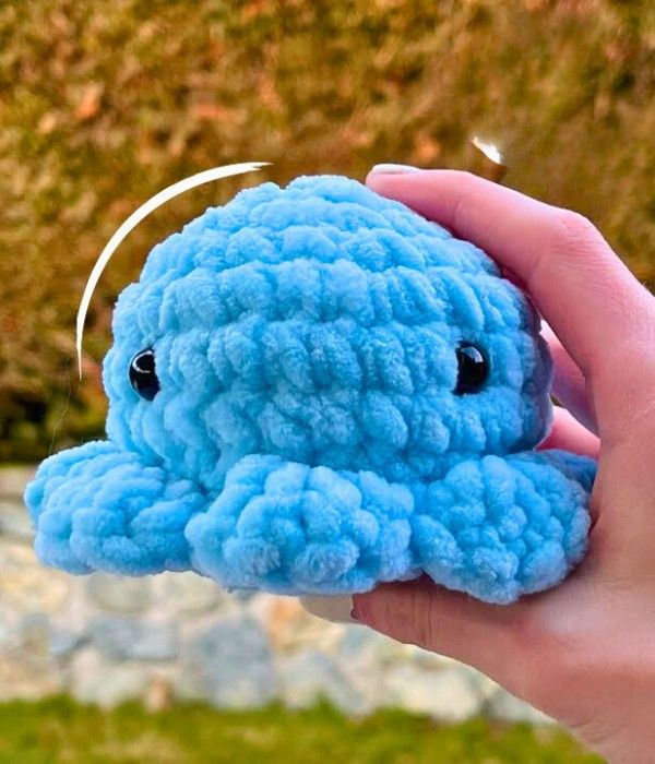 a hand holding a blue crocheted stuffed animal in front of a grassy area