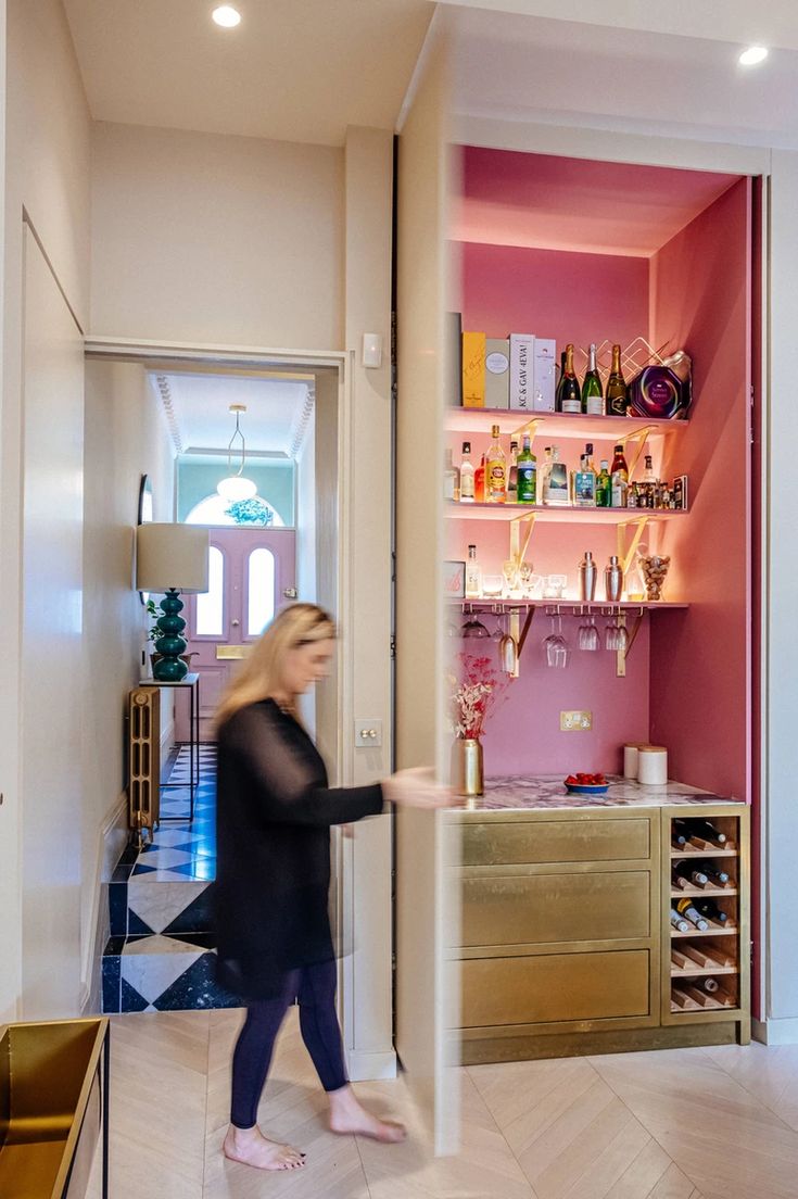 a woman walking through a room with pink walls and shelves filled with bottles on the wall