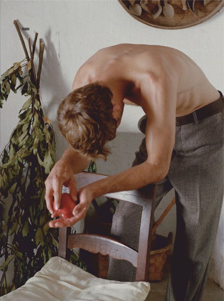 a shirtless man leaning on a chair with an apple in his hand