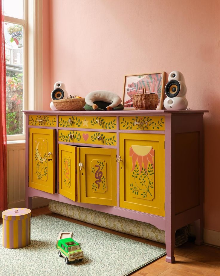 a pink and yellow cabinet with pictures on it next to a green toy car in front of a window