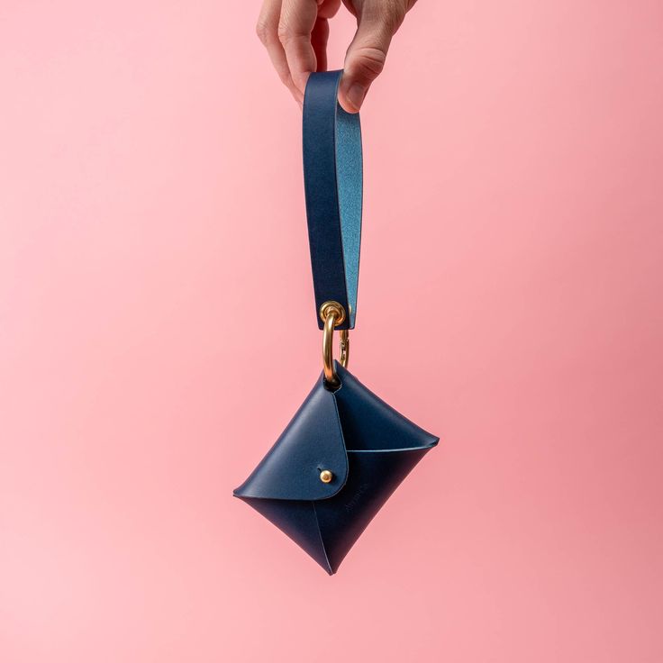 a hand holding a blue leather wallet on a pink background with the strap down to it's pocket