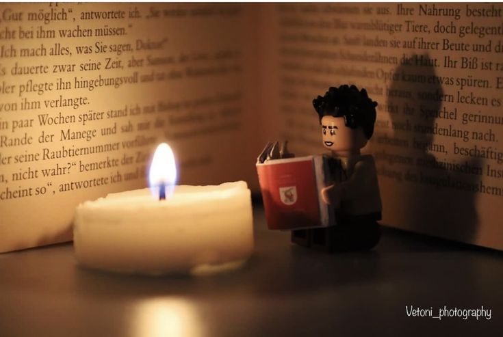 a lego man reading a book next to a lit candle on a table with an open book in the background