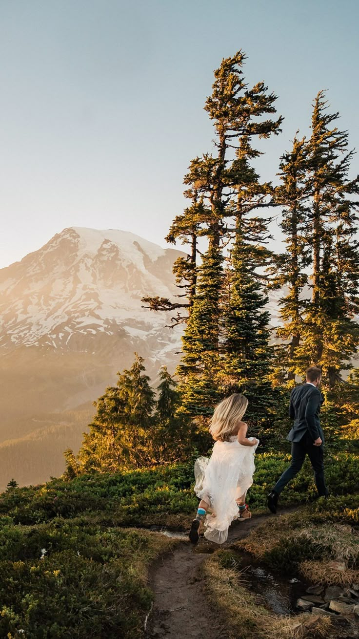 a man and woman walking down a trail in the mountains with snow capped mountain behind them