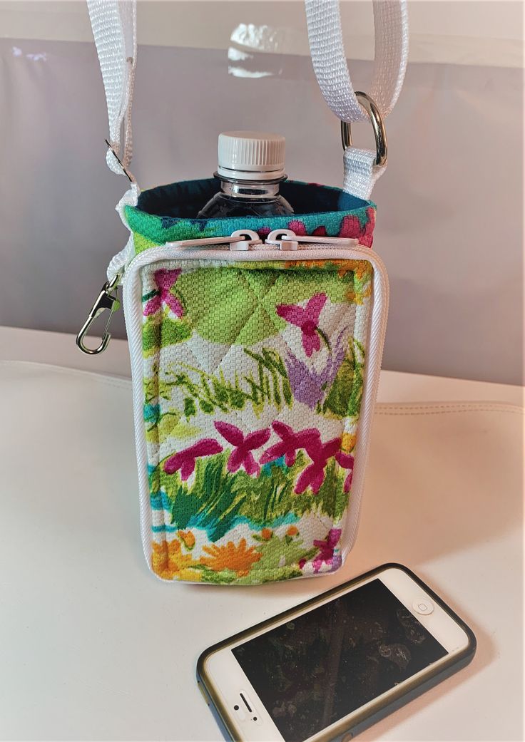 "Have you ever wished for a hands free way to carry around your water or soda bottle?   This holder is insulated to keep your water cool.  It has an adjustable strap to allow you to carry it off your shoulder or crossed over your chest.  The bottle holder has a zipper pocket for your phone or eyeglasses.  Inside the pocket there are pockets for cash or credit cards.    It also has a spring clip for your keys. This bottle holder was made from white cotton fabric with all over bright flowers.  The Drinks Bottle, Water Bottle Carrier, Soda Bottle, White Cotton Fabric, Bottle Carrier, Insulated Bags, Soda Bottles, Water Bottle Holders, Water Cooler