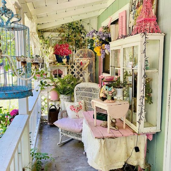 a porch filled with lots of plants and flowers next to a birdcage on top of a table