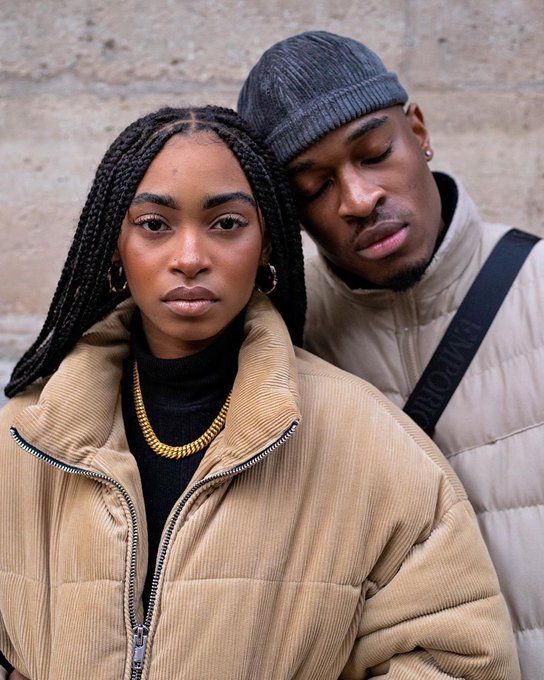 a man and woman standing next to each other in front of a stone wall wearing winter clothes