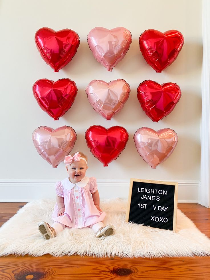 a baby sitting on a rug in front of heart shaped balloons and a sign that says valentine's day