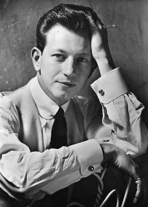 black and white photograph of a man sitting down with his hands on his head, wearing a tie