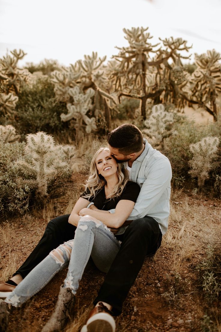 a man and woman sitting on the ground in front of cacti with their arms around each other