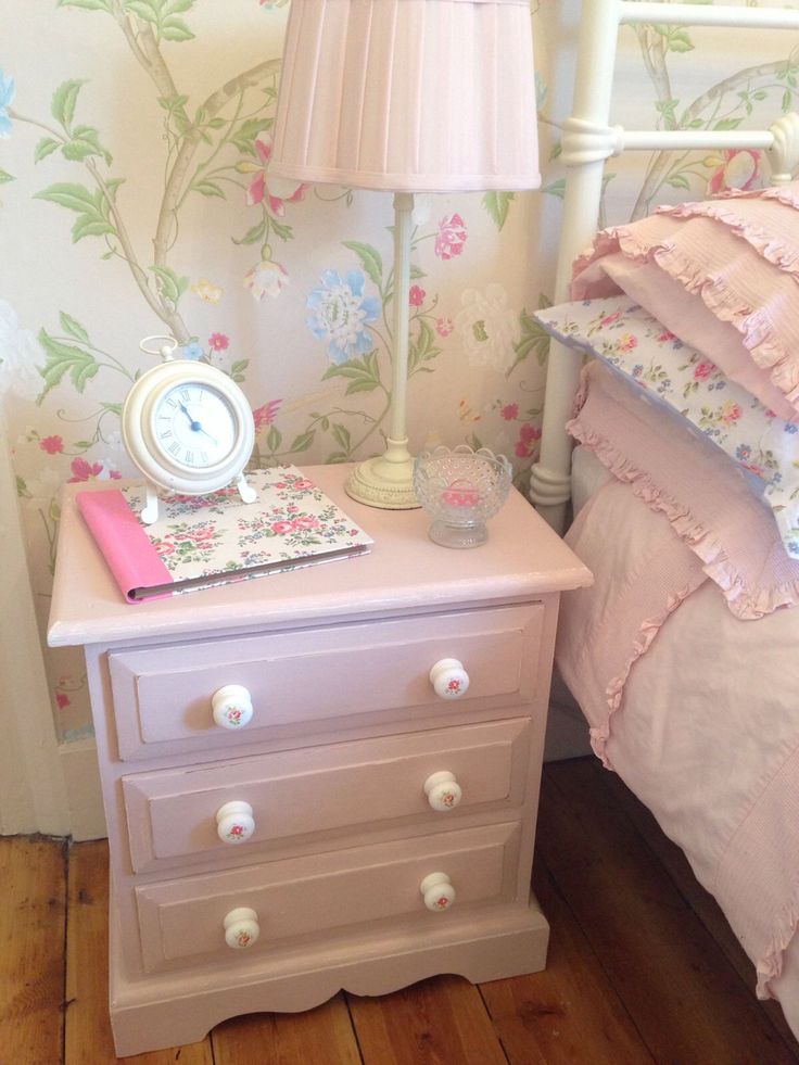 a pink nightstand with a clock and lamp on it next to a flowered wallpaper