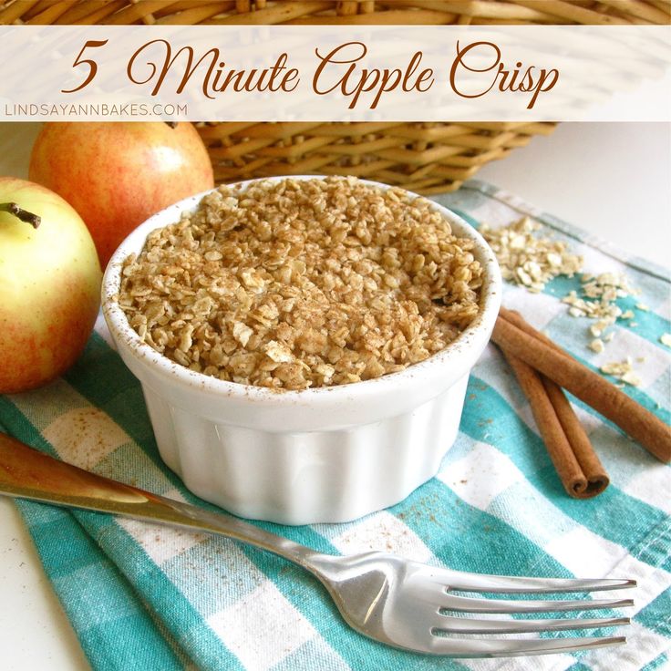 an apple crisp in a white bowl next to apples and cinnamon sticks on a blue checkered table cloth