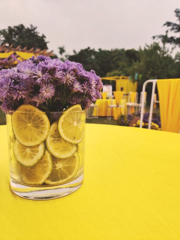 a glass vase filled with lemon slices and purple flowers on top of a yellow table