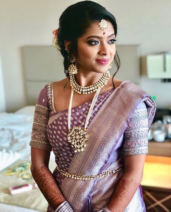 a woman in a purple and white sari with gold jewelry on her neck, standing next to a bed