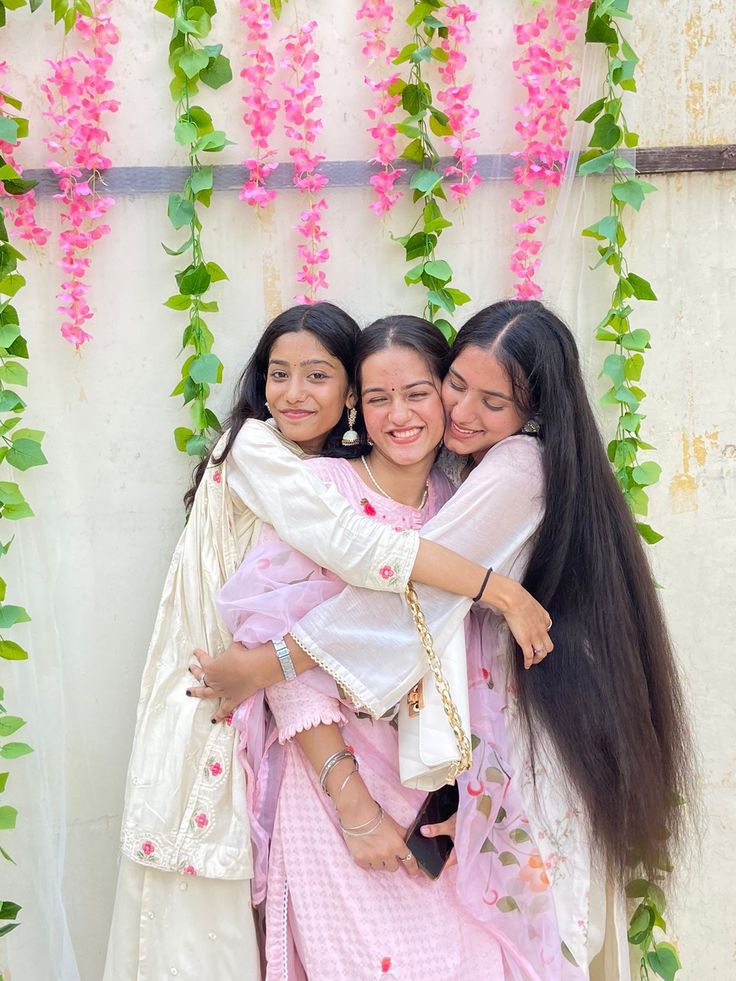 three young women hugging each other in front of a wall covered with pink flowers and greenery