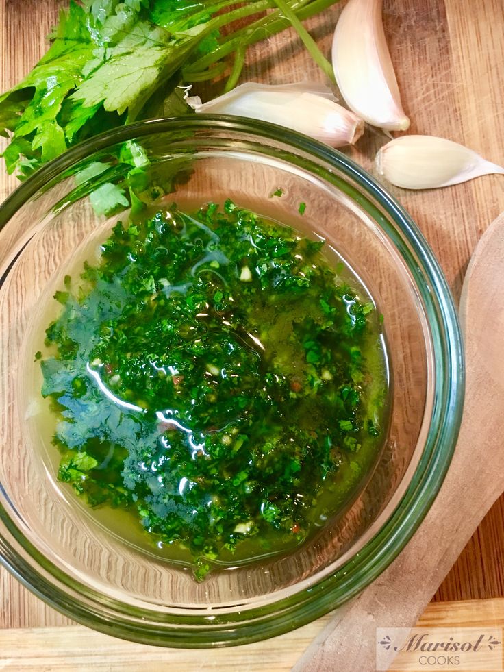 a glass bowl filled with green vegetables on top of a wooden cutting board next to garlic and parsley