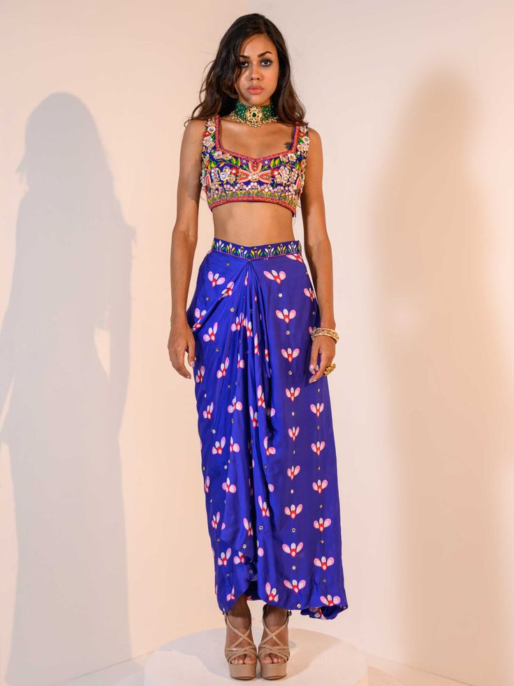 A three-piece Blue Indica Mirror Dhoti Skirt Set Paired with Bralette and Cape from the Juhi Bengani collection – Rang-Festive 2022. This gorgeous blue indica silk mirror dhoti skirt is paired with a heavy embroidered multicolored bralette blouse. The bright and vibrant colors with floral print, heavy embroidery of mirror, beads, stones, and thread sequence work enhance this satin dhoti skirt set. The indica blue dhoti skirt has a side opening at the waistline. This outfit is completed with a mu Blue Silk Sets For Diwali, Blue Silk Set For Diwali, Blue Palazzo Set For Reception And Festivals, Blue Silk Set For Navratri, Blue Festive Palazzo Set For Reception, Blue Fitted Palazzo Set With Traditional Drape, Royal Blue Designer Sharara, Bollywood Blue Sleeveless Sets, Blue Palazzo Set With Unstitched Blouse For Diwali