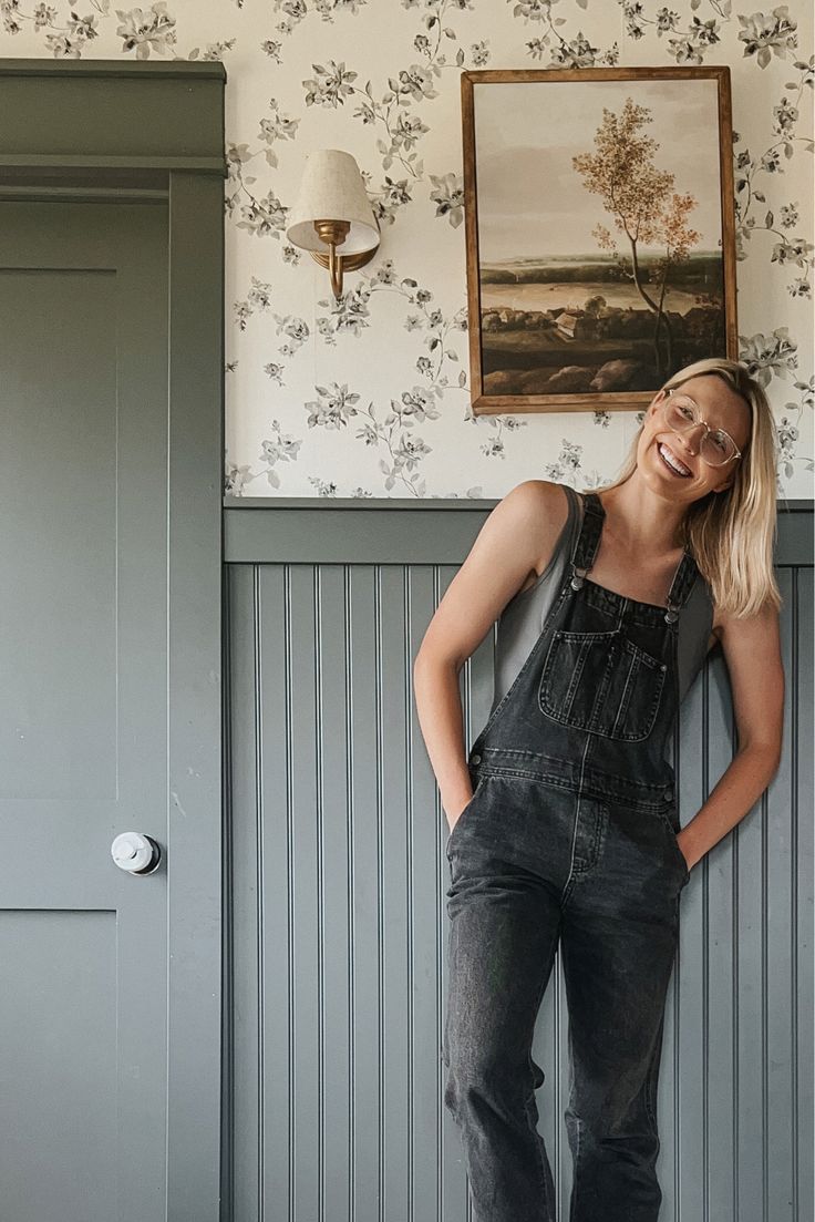 a woman in overalls leaning against a wall