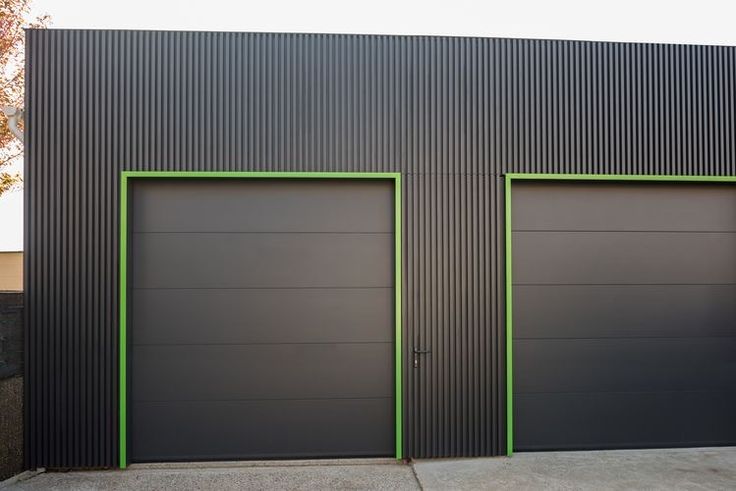 two gray garage doors with green trim in front of a black and grey building on a street