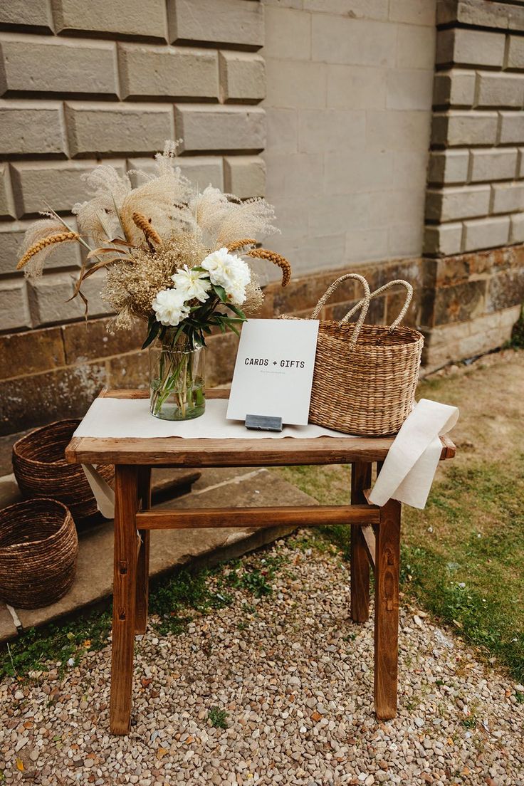 Wooden table and with flowers in glass vase and wicker basket with sign reading 'Cards Gifts' Picnic Basket Wedding Card Holder, Wicker Card Box Wedding, Gift Table Flowers Wedding, Boho Wedding Signing Table, Cards Gifts Table Wedding, Basket Card Box Wedding, Dried Flowers Wedding Decorations, Wooden Table Signs For Wedding, Boho Guest Sign In Table