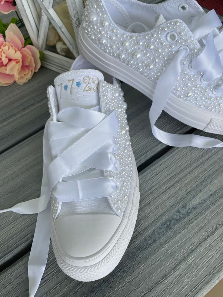 two white shoes with bows and pearls on them sitting on a wooden bench next to flowers