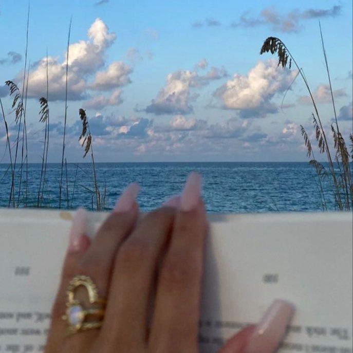 a woman's hand holding an open book with the ocean in the background