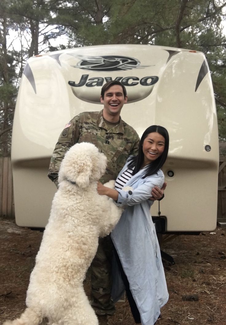 a man and woman standing next to a white dog in front of a camper