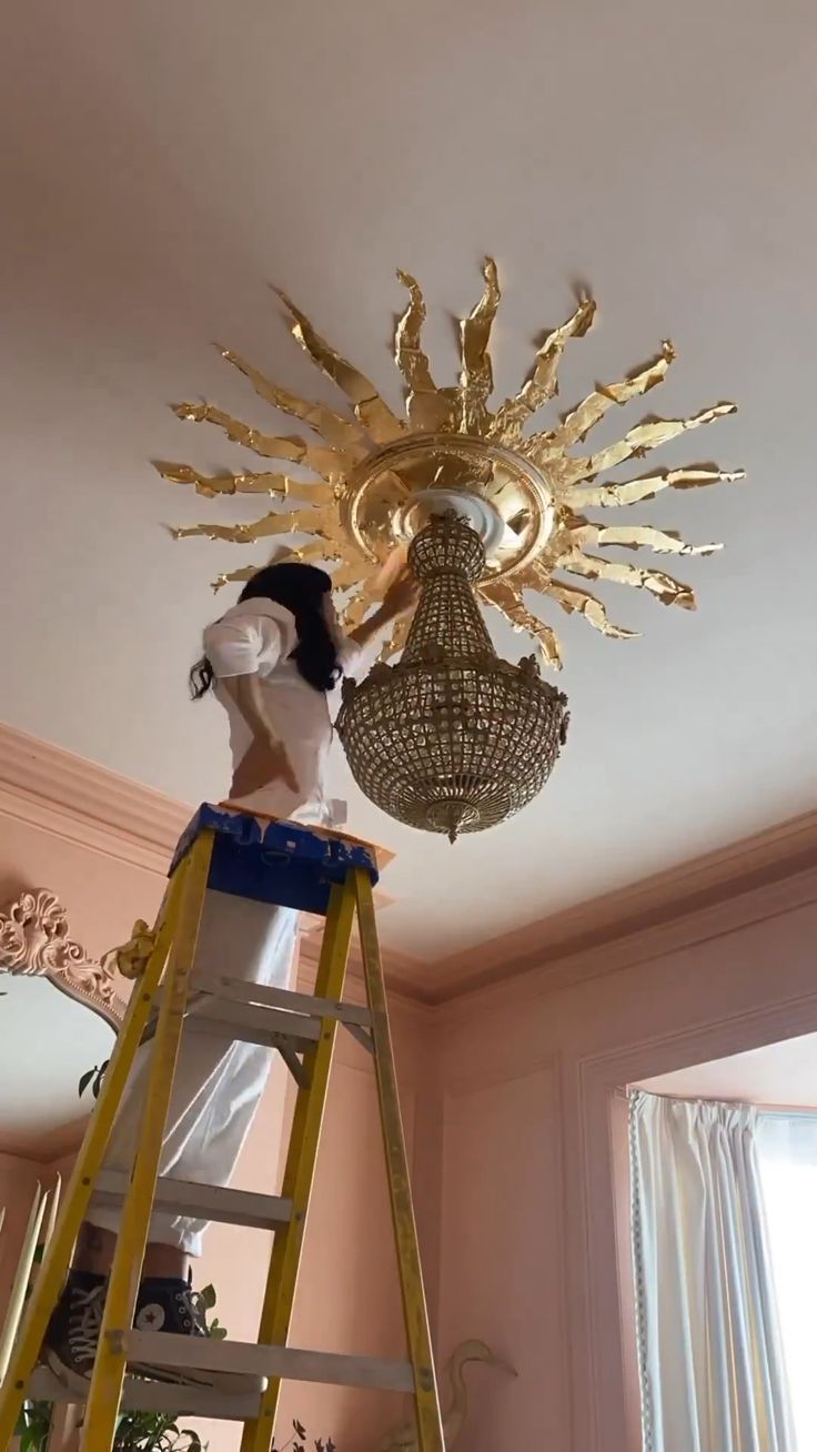 a gold sunburst light fixture being installed on a ladder in a pink room