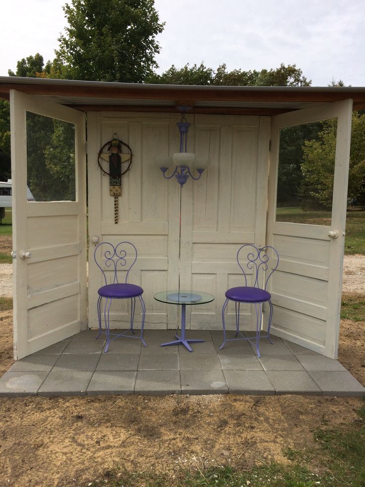 two purple chairs sitting at a table in front of a white shed with an open door